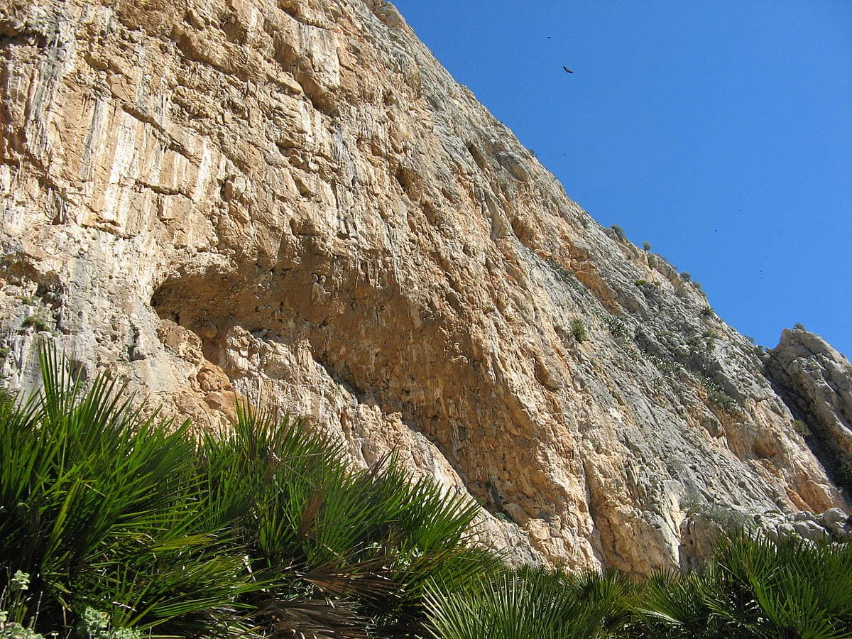 The Poema de Roca cave at the foot of the Frontales wall, seen from the path below. The written approach notes this cave is visible in the background from the start of Estrella Polar.