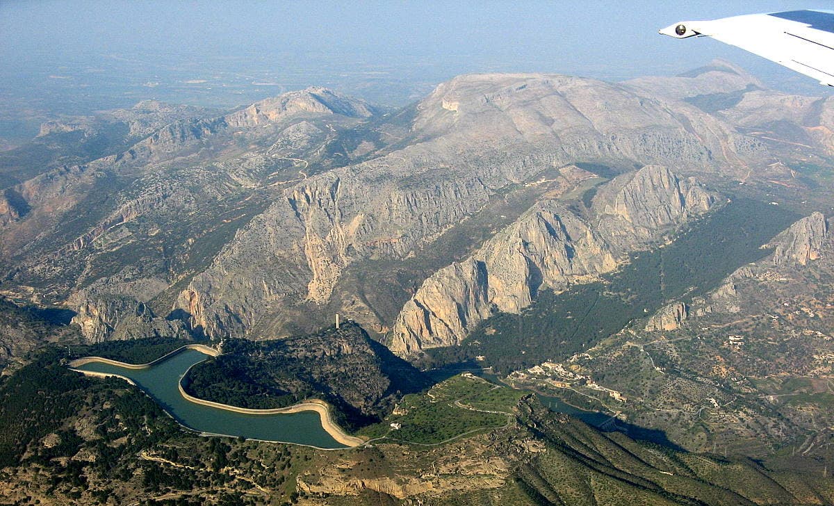 El Chorro from the air: the village and the lower reservoir are at bottom right, the Gaitanes gorge is the cleft left of centre, and the Frontales are the sunlit south-facing walls rising above the pine forest between the two. The upper car park lies between the village and the wall.