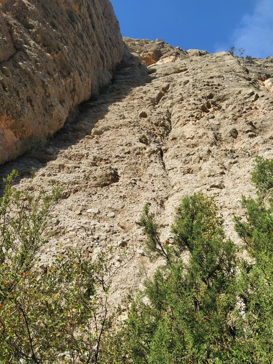 Looking up from the start of Tanger (September 2023): the conglomerate slab with the steeper wall forming a corner on its left, bushes at the base.