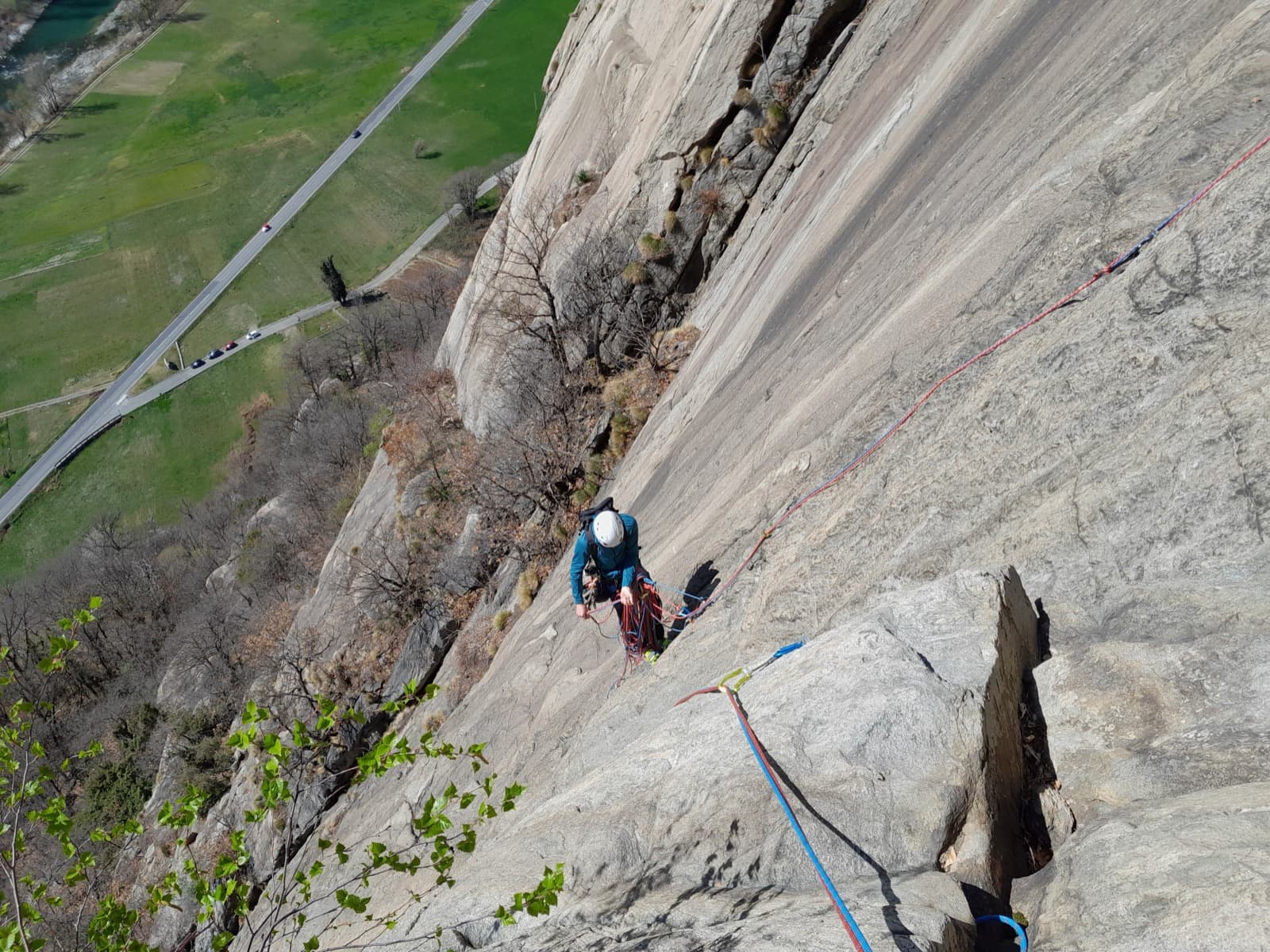 Looking down the gneiss slabs of Bucce d'Arancia to a climber at a belay, with the valley road and fields at the foot of the Paretone di Arnad below.