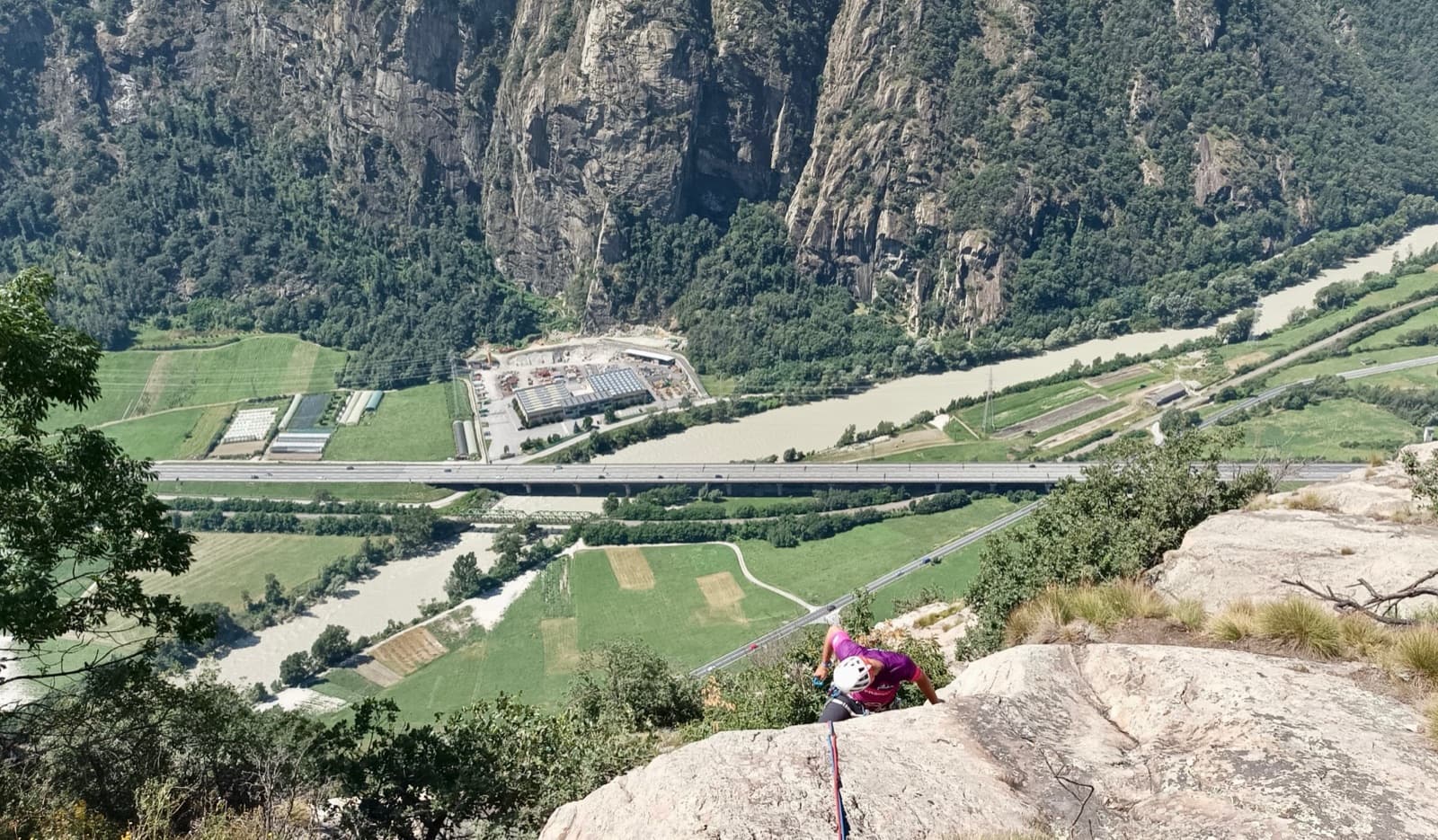 A climber arriving on a ledge high on Lo Dzerby, with the Dora Baltea, the motorway and the cliffs across the valley below.