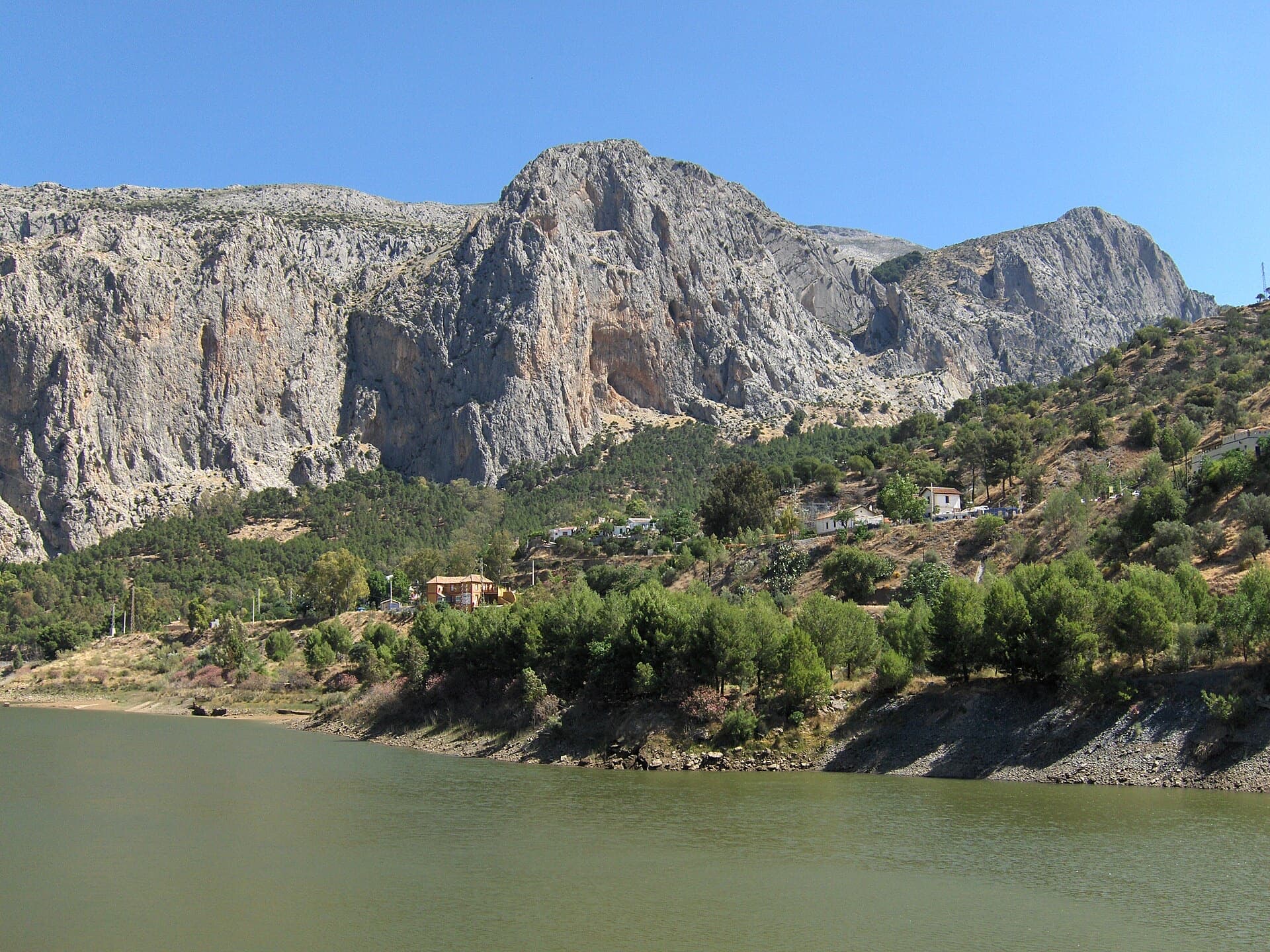 The Frontales cliffs above El Chorro seen across the Tajo de la Encantada reservoir, with the big Poema de Roca cave at the foot of the central dome. Apocalipsis is on this wall; the line is not marked.
