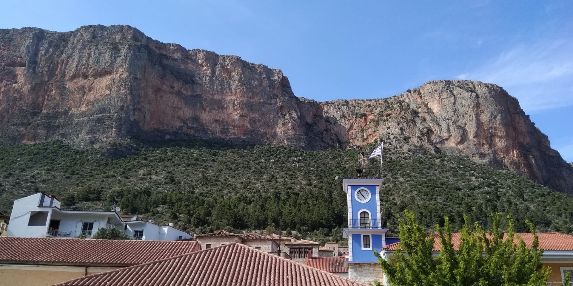 The red limestone wall of Kokkinovrachos rising directly above the rooftops and clock tower of Leonidio. Kapi Reborn is on the south-east face of this cliff; the exact line is not marked.